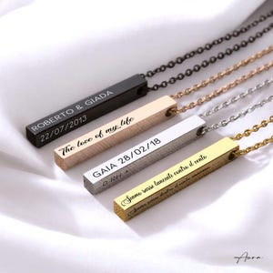 Può includere: Quattro collane a barra personalizzate in nero, oro, argento e oro rosa. Ogni ciondolo presenta testo inciso, inclusi nomi e date. Le collane sono appese a delicate catene, esposte su una superficie bianca.