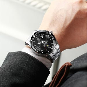 May include: A silver wristwatch with a black face is displayed on a wrist. The watch has a stainless steel band and a black dial with white numerals and hands. The watch is being worn with a white dress shirt and a dark suit jacket.
