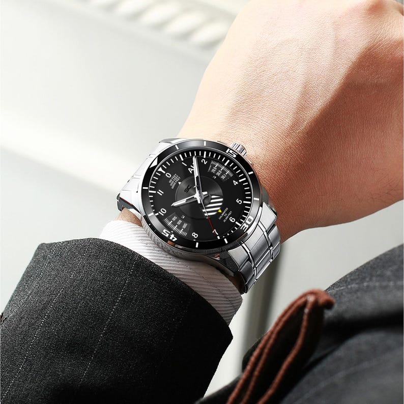 May include: A silver wristwatch with a black face is displayed on a wrist. The watch has a stainless steel band and a black dial with white numerals and hands. The watch is being worn with a white dress shirt and a dark suit jacket.