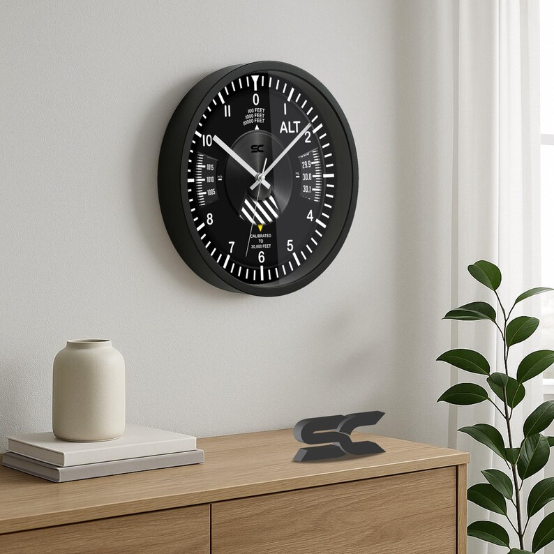 May include: A black wall clock with white markings and hands, resembling an aircraft altimeter. The clock face displays numbers and the word "ALT". The clock is mounted on a white wall. A small black sculpture sits on a wooden surface.