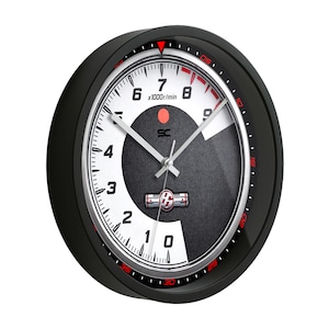 May include: Black and white wall clock with a speedometer-style face. The clock features a black bezel, a white and gray dial with red accents, and silver hands. The face includes the text "x1000r/min".