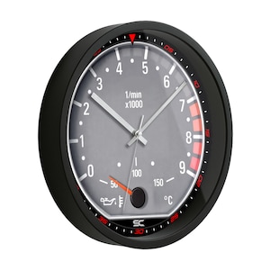 May include: Black and gray wall clock with a design resembling a car's tachometer. The clock face features numbers, markings, and gauges in red and white. The clock has a black bezel and hands.