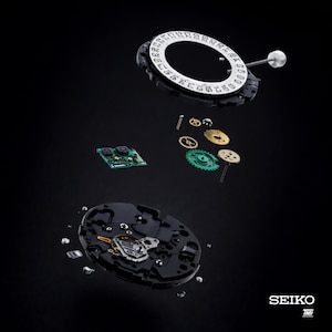 May include: An exploded view of a Seiko watch mechanism, showcasing its intricate components. The image features a black background with various parts suspended in mid-air, including gears, a circuit board, and the date dial. The Seiko logo is visible at the bottom right.