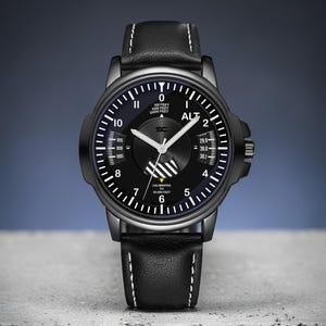 May include: Black wristwatch with a black dial and white markings. The watch features a black leather band with white stitching. The watch face includes the text "ALT" and other technical details. The watch is set against a gray and blue background.
