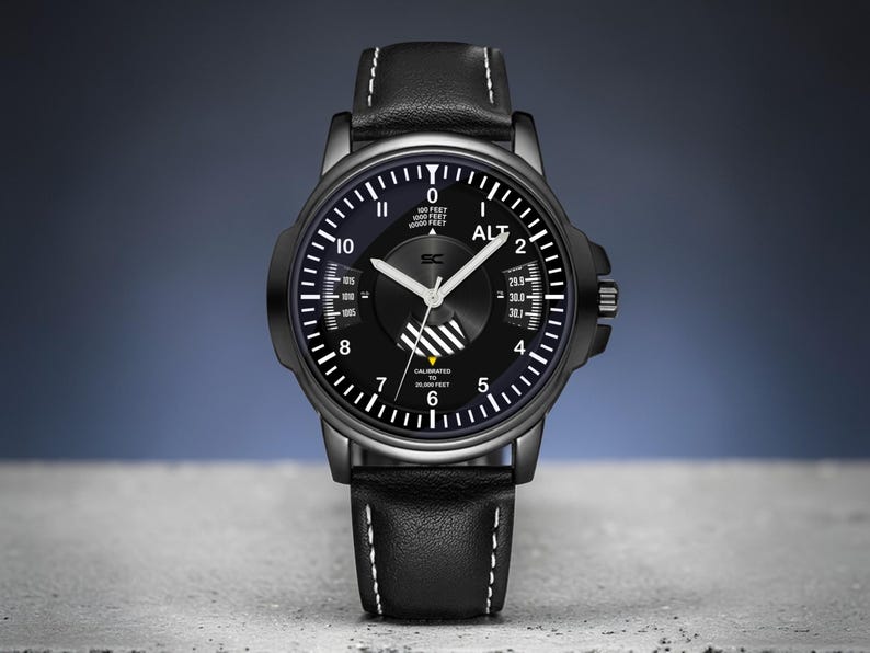 May include: Black wristwatch with a black dial and white markings. The watch features a black leather band with white stitching. The watch face includes the text "ALT" and other technical details. The watch is set against a gray and blue background.