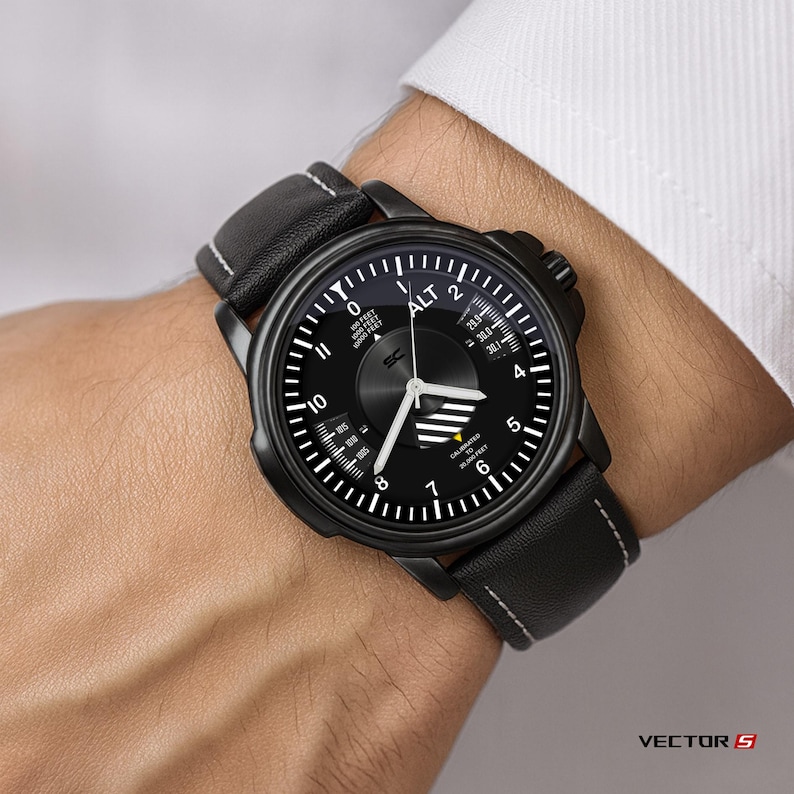 May include: Black wristwatch with a round face and a black leather band. The watch face features white markings and hands, with the word "ALT" at the top. The band has white stitching.