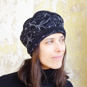 Peut inclure: Un bonnet noir avec un motif de dentelle florale argentée. Le chapeau est fait d'une matière douce et possède une large bande à la base. Le chapeau est porté par une personne.