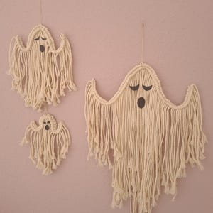 Könnte beinhalten: Drei Wandbehänge in Geisterform aus Makramee in verschiedenen Größen. Die Geister sind aus cremefarbenem Seil mit schwarzen Gesichtern gefertigt. Der größte Geist befindet sich in der Mitte, mit einem mittleren und kleinen Geist links. Sie hängen an einer rosa Wand.