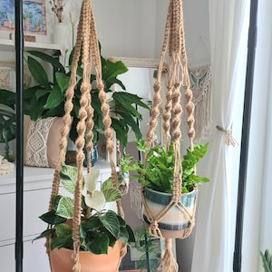 Op de afbeelding: Twee macramé plantenhangers met potplanten. Eén hanger houdt een plant in een oranje pot, de andere in een blauw-witte pot. De hangers zijn opgehangen aan metalen ringen en een zwarte metalen standaard.
