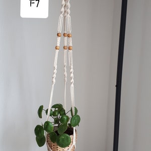 May include: A white macrame plant hanger with wooden beads hangs from a black metal pipe. A green plant in a woven basket is suspended from the hanger. The hanger is labeled "F7".