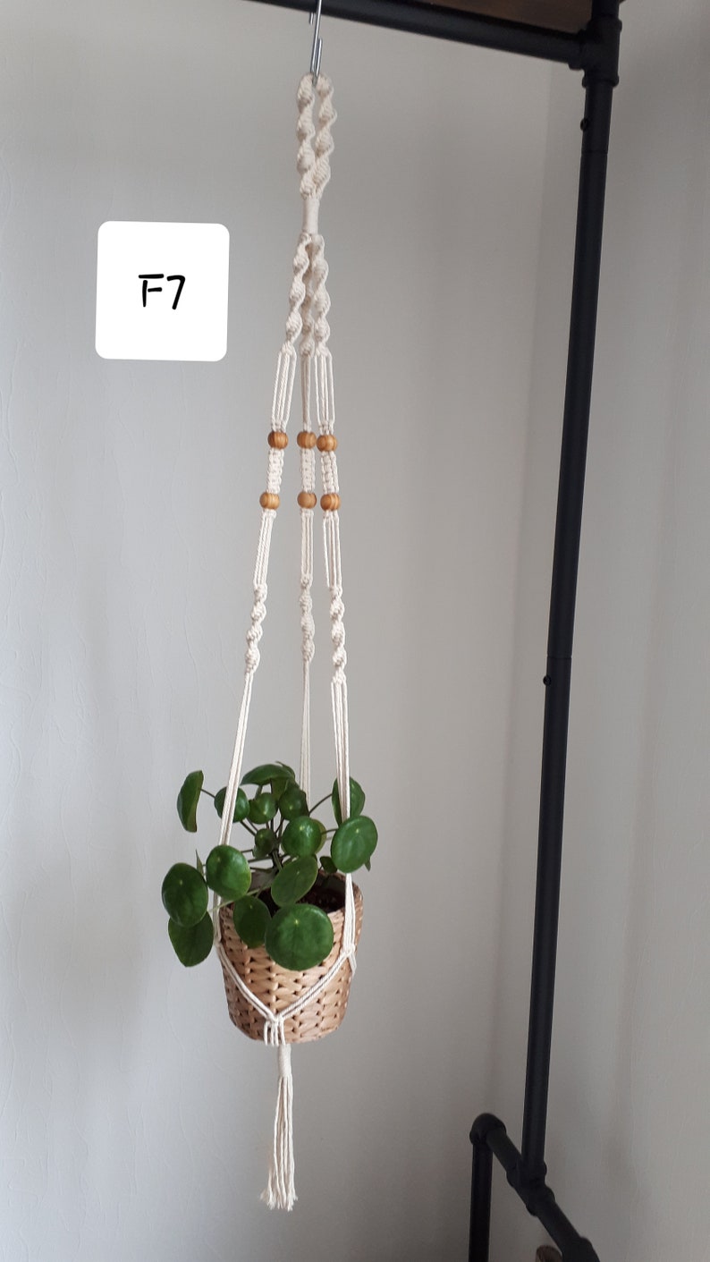 May include: A white macrame plant hanger with wooden beads hangs from a black metal pipe. A green plant in a woven basket is suspended from the hanger. The hanger is labeled "F7".