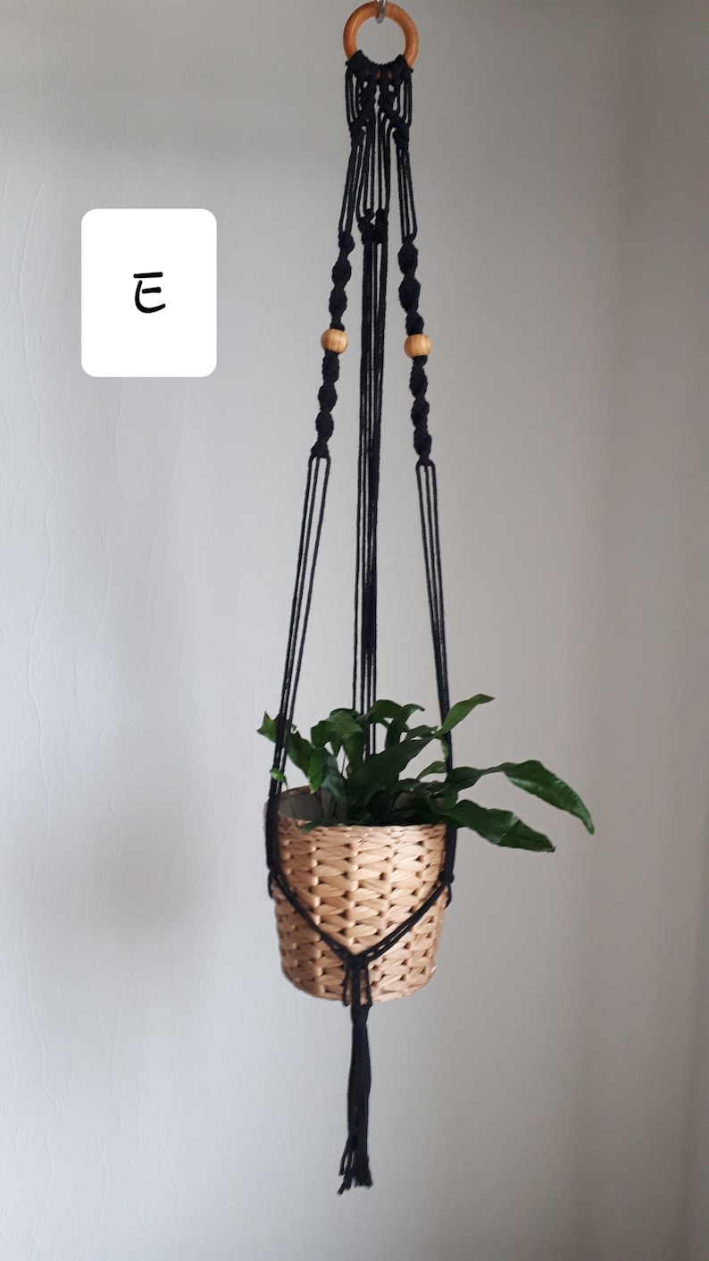 May include: Black macrame plant hanger with wooden beads and a wooden ring at the top. A small potted plant is hanging from the hanger.