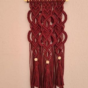 Könnte beinhalten: Ein burgunderfarbener Makramee-Wandbehang mit Holzleiste und Quasten. Der Wandbehang zeigt ein sich wiederholendes Muster aus Knoten und ist mit Holzperlen verziert.