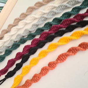 Könnte beinhalten: Eine Sammlung farbenfroher, verdrehter Garnstränge in verschiedenen Farbtönen, darunter Beige, Weiß, Grau, Grün, Burgunderrot, Schwarz, Gelb und Orange. Die Stränge sind diagonal angeordnet und zeigen ihr strukturiertes, seilartiges Aussehen. Das Bild ist gut beleuchtet und hebt die lebendigen Farben hervor.