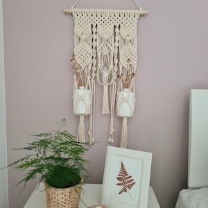 Könnte beinhalten: Ein Makramee-Wandbehang mit drei weißen Keramikvasen mit Trockenblumen. Ein gerahmter Farn-Druck, ein Topf mit Farn und eine Geode stehen auf einem weißen Tisch. Die Wand ist helllila gestrichen.