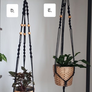 May include: Two black macrame plant hangers with wooden beads. The hangers are hanging from a black metal rack. One hanger has a green plant in a woven basket, and the other has a leafy plant in a woven basket.