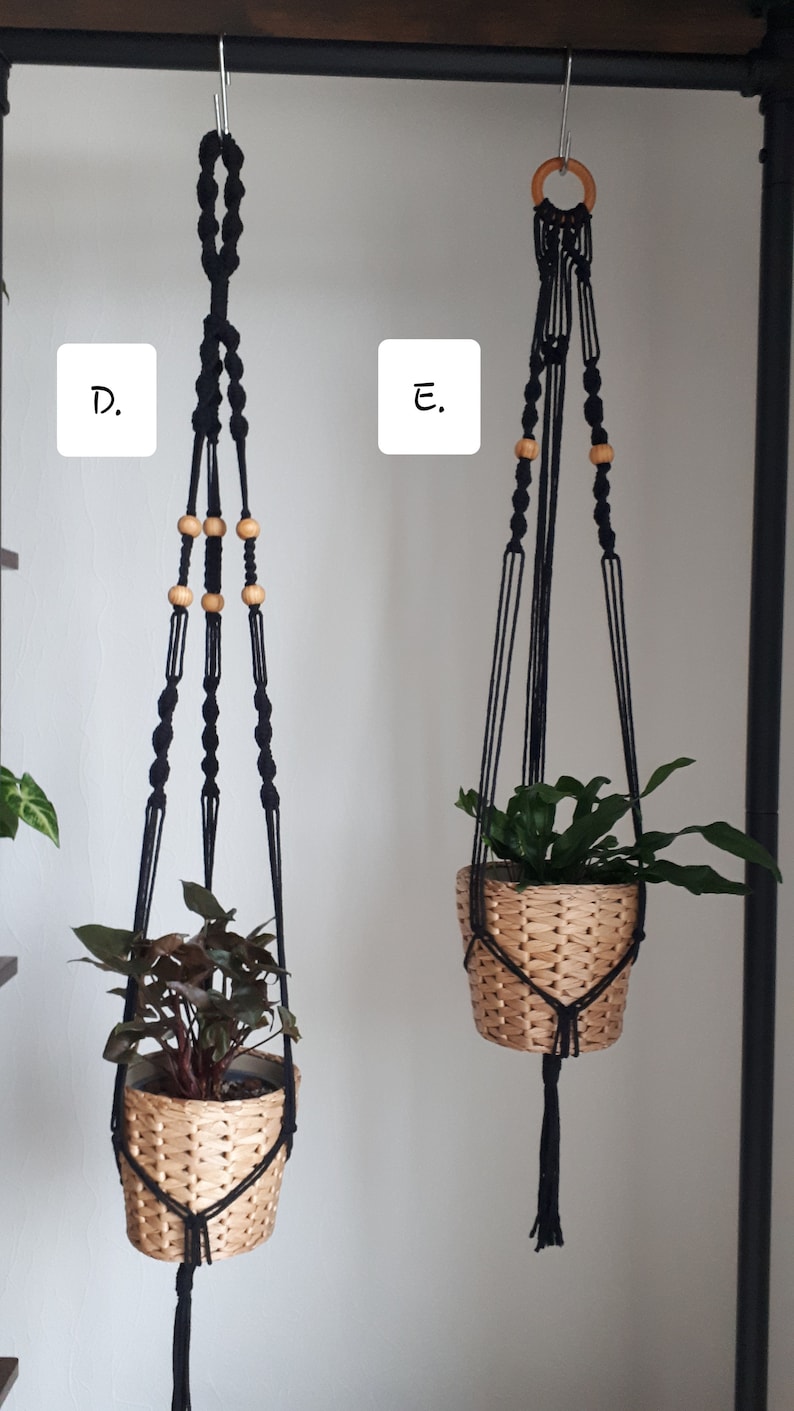 May include: Two black macrame plant hangers with wooden beads. The hangers are hanging from a black metal rack. One hanger has a green plant in a woven basket, and the other has a leafy plant in a woven basket.