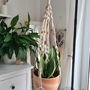 Op de afbeelding: Een macramé plantenbak met een terracotta pot met een sanseveria. De gebroken witte katoenen hanger heeft houten kralen en een houten ring aan de bovenkant. De plant heeft groene en gele bladeren. De hanger hangt aan een zwarte metalen standaard.