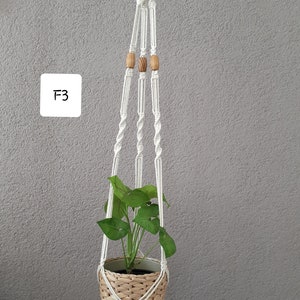May include: White macrame plant hanger with wooden beads, holding a potted plant with green leaves. The hanger is hanging from a hook on a gray wall. The text "F3" is written on a white square in the upper left corner of the image.