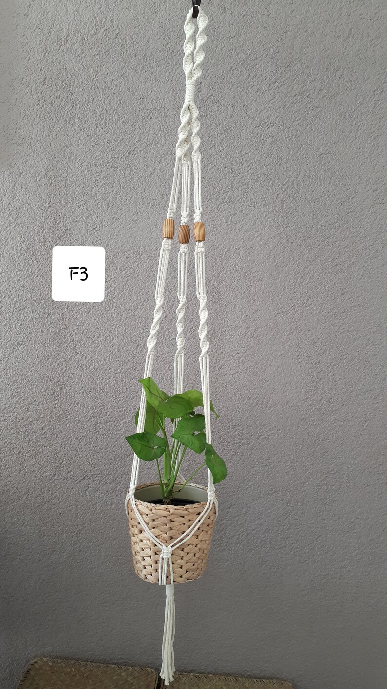 May include: White macrame plant hanger with wooden beads, holding a potted plant with green leaves. The hanger is hanging from a hook on a gray wall. The text "F3" is written on a white square in the upper left corner of the image.