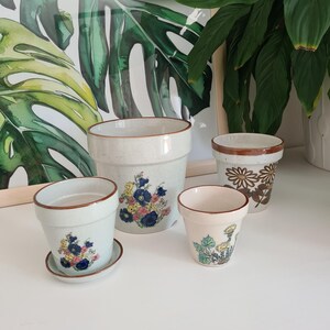Könnte beinhalten: Eine Sammlung von vier Keramik-Pflanztöpfen mit gesprenkelten cremeweißen Oberflächen und braunen Rändern. Zwei Töpfe zeigen bunte Blumenmotive, einer hat ein braunes Gänseblümchenmuster und ein kleiner Topf enthält einen passenden Untersetzer.