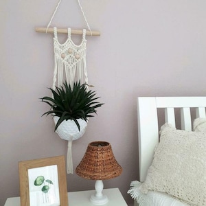 Op de afbeelding: Een witte macramé plantenhanger met houten lat en drie houten kralen hangt aan een lichtroze muur. Een groene succulent plant staat in een witte keramische pot. Een wit nachtkastje met een bruine rieten lampenkap en een ingelijst aquarelprint van een groene plant bevindt zich op de voorgrond.
