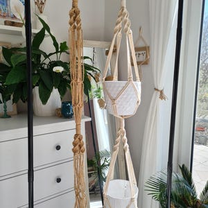 Op de afbeelding: Twee macramé plantenhangers met witte potten die aan een zwart metalen frame hangen. De ene is een lang, verticaal ontwerp in beige, terwijl de andere twee witte potten met een ruitpatroon ondersteunt. De kamer heeft een heldere, luchtige uitstraling.