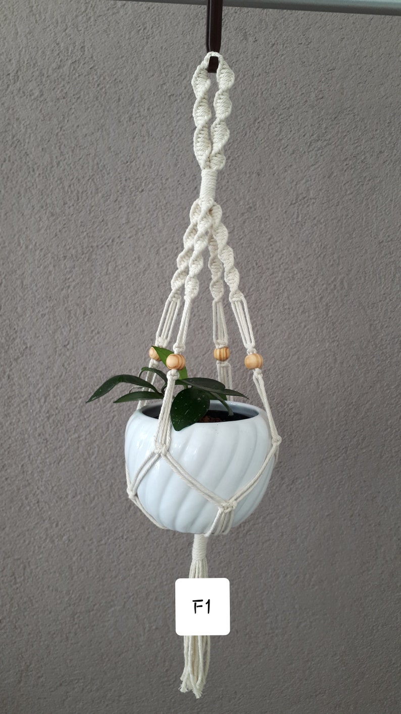 May include: White ceramic planter with a green plant inside, hanging from a macrame plant hanger with wooden beads. The hanger has a long tassel at the bottom. The text "F1" is written on a white tag attached to the tassel.