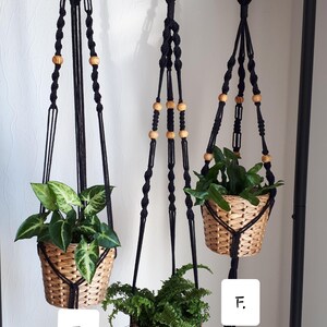 May include: Three black macrame plant hangers with wooden beads hanging from a black metal rod. Each hanger has a potted plant in a woven basket. The hangers are labeled D, E, and F.