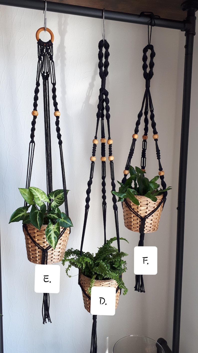 May include: Three black macrame plant hangers with wooden beads hanging from a black metal rod. Each hanger has a potted plant in a woven basket. The hangers are labeled D, E, and F.