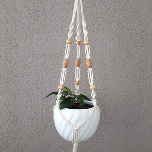 May include: White macrame plant hanger with wooden beads and a white ceramic pot with a green plant inside. The hanger has a tassel at the bottom and the label "F2" is visible.