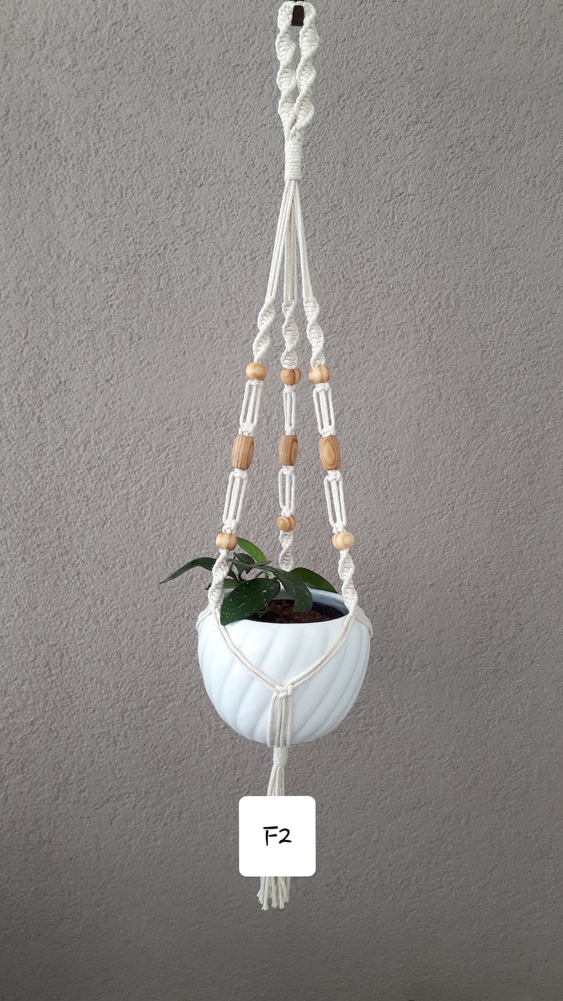 May include: White macrame plant hanger with wooden beads and a white ceramic pot with a green plant inside. The hanger has a tassel at the bottom and the label "F2" is visible.