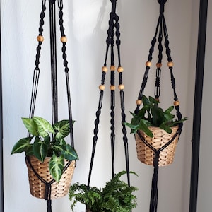 May include: Three black macrame plant hangers with wooden beads, each holding a potted plant. The plants are a fern, a leafy green plant, and a variegated plant.