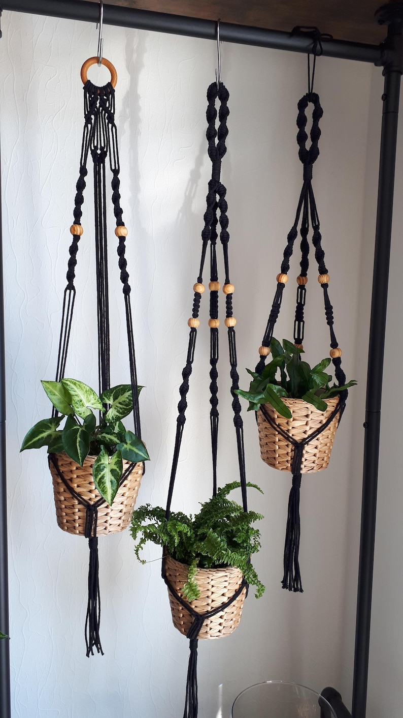 May include: Three black macrame plant hangers with wooden beads, each holding a potted plant. The plants are a fern, a leafy green plant, and a variegated plant.