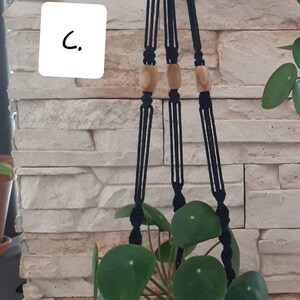 May include: A black macrame plant hanger with wooden beads hangs from a white brick wall. A green plant with round leaves is in a woven basket.