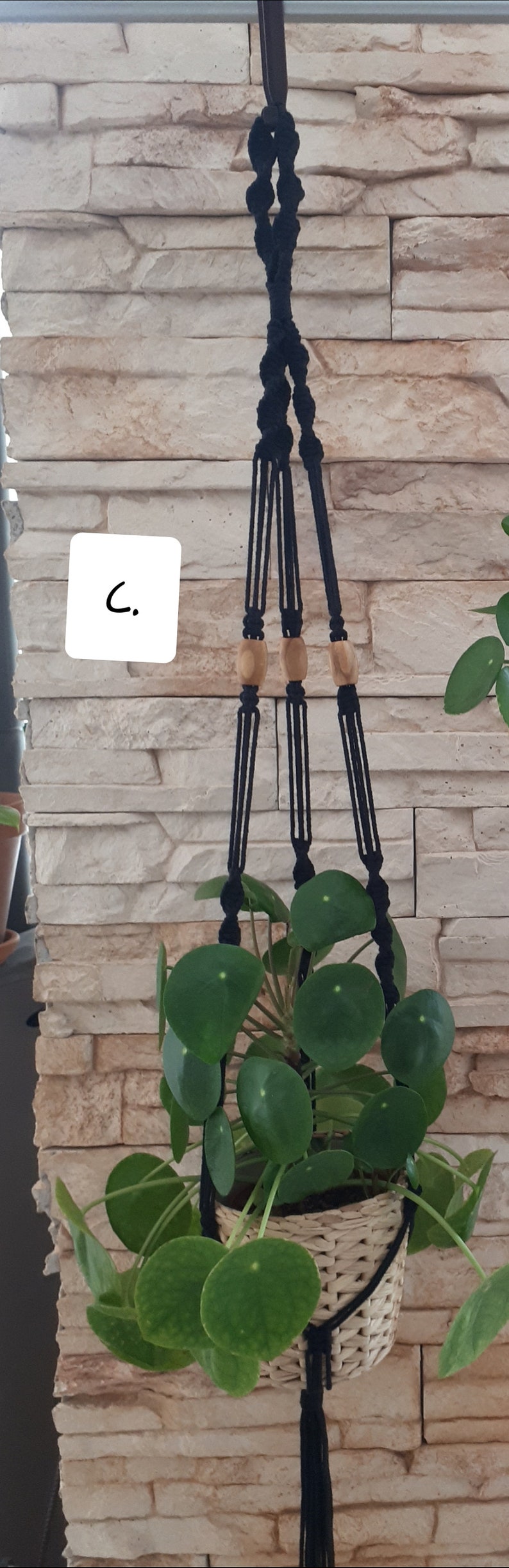 May include: A black macrame plant hanger with wooden beads hangs from a white brick wall. A green plant with round leaves is in a woven basket.