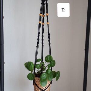 May include: Black macrame plant hanger with wooden beads, holding a potted plant with green leaves. The hanger is hanging from a hook on a white wall. The text "D." is written on a white square.