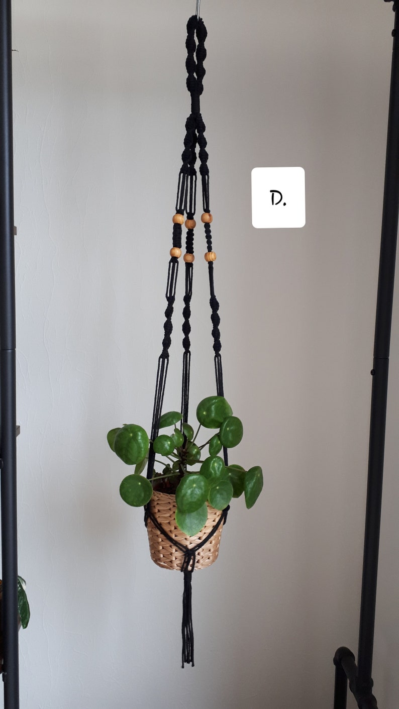 May include: Black macrame plant hanger with wooden beads, holding a potted plant with green leaves. The hanger is hanging from a hook on a white wall. The text "D." is written on a white square.