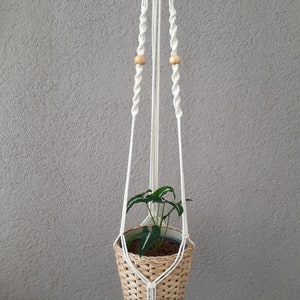 May include: White macrame plant hanger with wooden beads and a woven basket pot with a green plant inside. The hanger has a wooden ring at the top and a label that says "F4".
