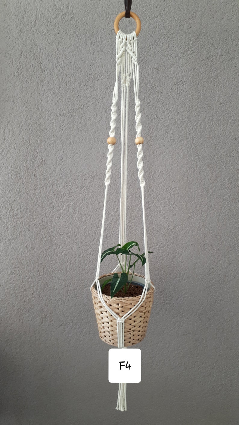 May include: White macrame plant hanger with wooden beads and a woven basket pot with a green plant inside. The hanger has a wooden ring at the top and a label that says "F4".