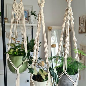 Op de afbeelding: Drie macramé plantenhangers met houten ringen en kwastjes, elk met een andere potplant. De potten zijn in tinten groen, wit en grijs. De hangers zijn opgehangen aan een zwarte metalen stang, wat een bohemien esthetiek creëert.