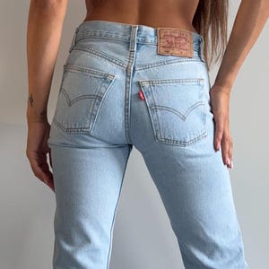 May include: Light blue denim jeans featuring a high-rise waist, two back pockets with signature arcuate stitching, a small red tab on the right pocket, and a tan leather patch on the waistband. The jeans have a straight-leg cut and a classic five-pocket design.