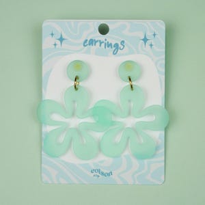 May include: Mint green flower-shaped earrings with gold-colored hooks. The earrings are displayed on a light blue card with the word "earrings" and a wavy pattern. The brand name "colson" is printed at the bottom of the card.