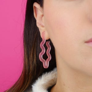 Könnte beinhalten: Ein Paar hellrosa, wellenförmige Ohrringe. Die Ohrringe haben ein einzigartiges, abstraktes Design mit einem glatten, durchscheinenden Aussehen. Die Ohrringe werden am Ohr einer Person getragen, vor einem hellrosa Hintergrund.
