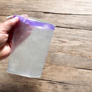 May include: A white mesh laundry bag with a purple trim and a white zipper is held by a hand. The bag is partially filled. The background is a wooden surface.
