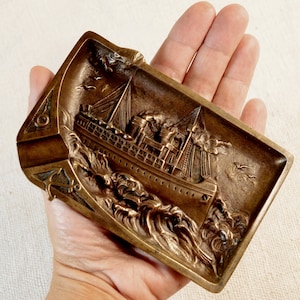May include: A vintage brass ashtray with a detailed relief of a ship sailing on the waves. The ashtray has a rectangular shape with rounded corners and a raised edge. The ship is depicted in the center of the ashtray, with its sails billowing in the wind. The waves are sculpted in a realistic manner, with foam crests and swirling water.