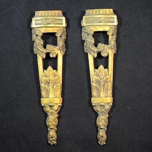 Puede incluir: Dos elementos arquitectónicos antiguos de latón ornamentado. Cada pieza presenta una parte superior rectangular con un diseño decorativo, una abertura cuadrada central y intrincadas tallas de hojas y flores. Las piezas miden aproximadamente 25 cm de largo.