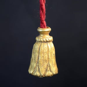 Op de afbeelding: Een goudkleurige, klokvormige decoratieve trekker met een getextureerd oppervlak en een rood touw. De trekker heeft een gedetailleerd ontwerp en hangt tegen een donkere achtergrond. Het touw is dieprood.