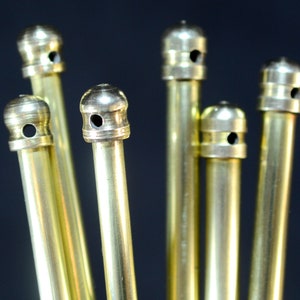 Può includere: Primo piano di diverse aste in ottone con cime decorative e arrotondate. Ogni asta ha un piccolo foro vicino alla sommità. Le aste sono disposte su uno sfondo scuro, con l'ottone che riflette la luce.
