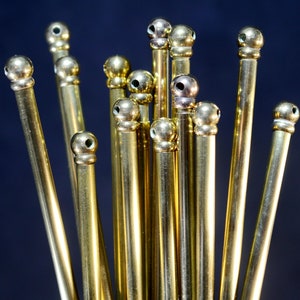 Könnte beinhalten: Eine Gruppe von 13 goldfarbenen Metallstäben mit abgerundeten Köpfen. Die Stäbe sind in einem Cluster angeordnet, wobei einige höher sind als andere.